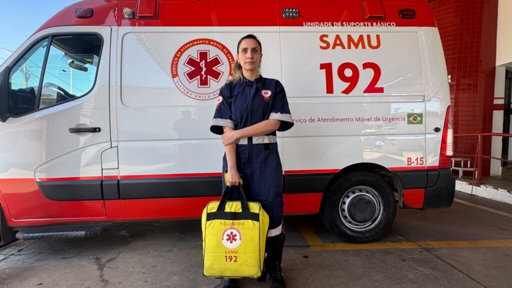 Fotografia de corpo inteiro da médica psiquiatra Bruna Rossi posicionada em pé, com os braços cruzados, em frente a uma ambulância do SAMU (Unidade de Suporte Básico, identificada como "B-15"). Ela veste o uniforme azul da corporação e segura à sua frente uma mala de atendimento amarela com inscrições voltadas para a especialidade de psiquiatria e o número 192.