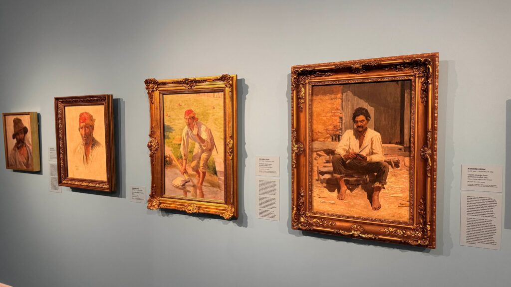 Várias pinturas emolduradas com molduras douradas e acompanhadas por fichas informativas encontram-se expostas numa parede de tom azul claro de uma galeria ou museu.