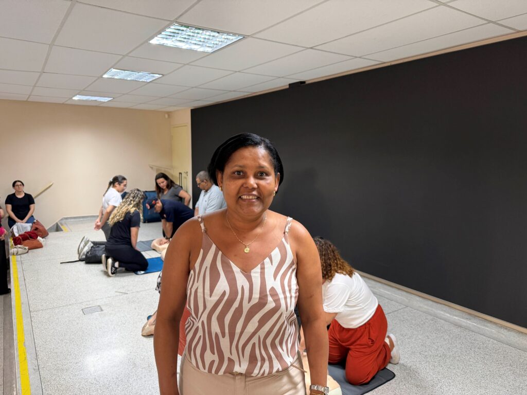 Retrato em plano médio da professora Elizabete Maria Câmara sorrindo para a câmara, com outros participantes a praticar manobras ao fundo no chão do local.