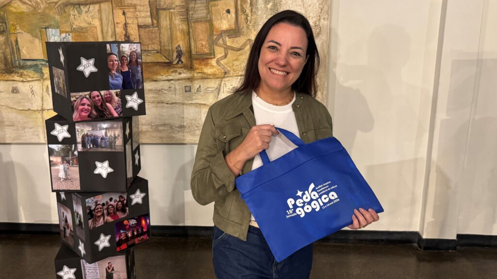 A professora Vanessa Abdala Guarda sorri enquanto segura a bolsa oficial da 18.ª Jornada Pedagógica ao lado de uma estrutura decorativa de cubos fotográficos.