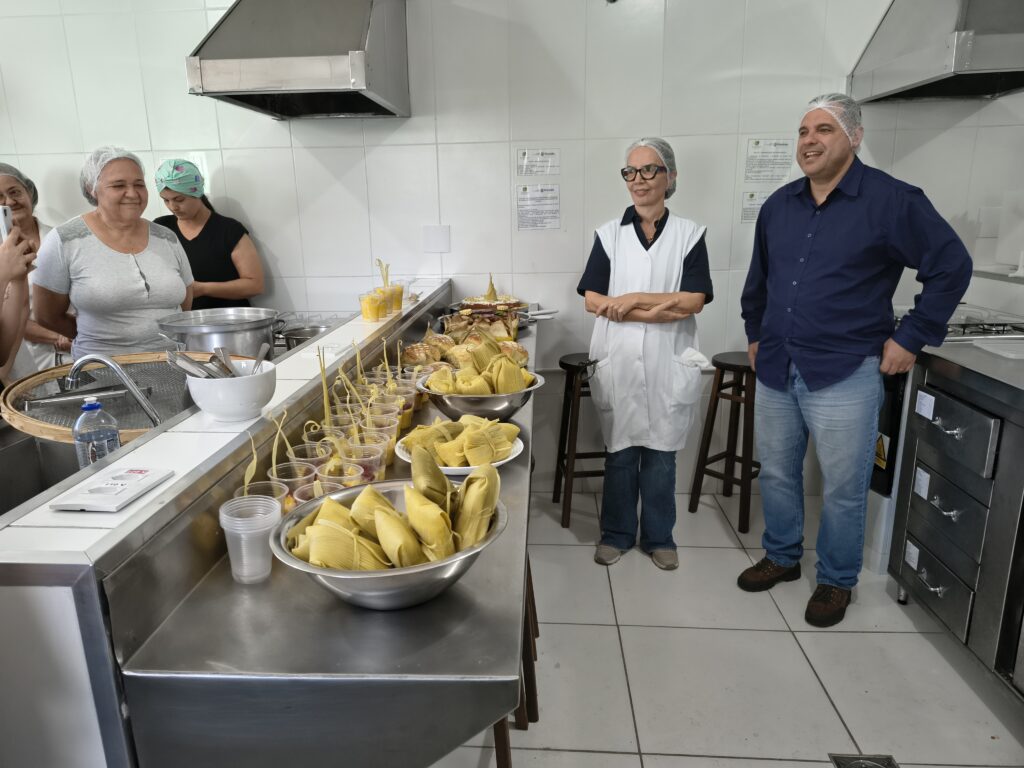 Participantes e instrutores de touca protetora reunidos numa cozinha industrial, ao lado de um balcão em inox repleto de alimentos preparados à base de milho, como pamonhas e sobremesas em copos.
