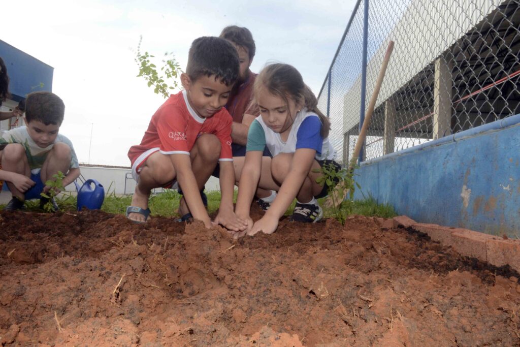 Duas crianças agachadas num canteiro delimitado por tijolos mexem na terra com as mãos para fixar uma muda, aprendendo na prática os cuidados com o solo e o cultivo.