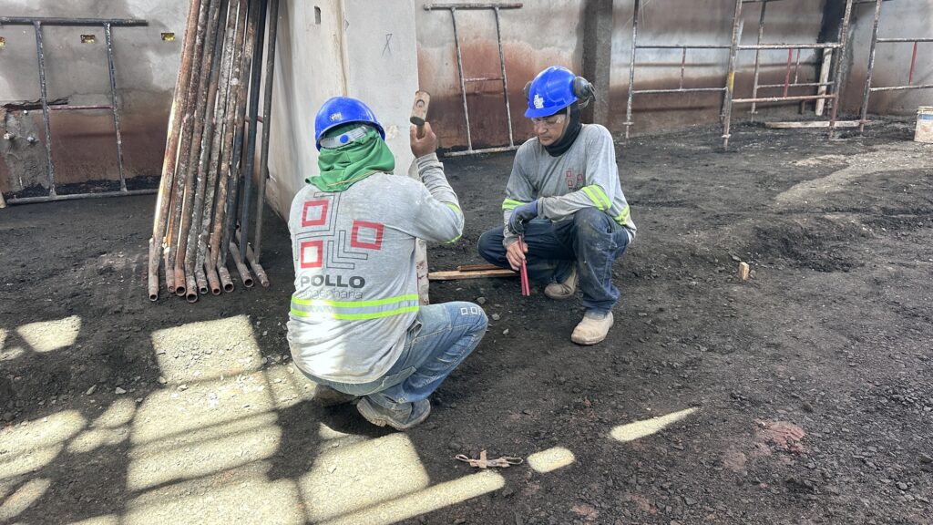 Dois trabalhadores da construção civil com capacete azul e equipamento de proteção trabalham agachados no chão de terra no interior em obras do Mercado Municipal.