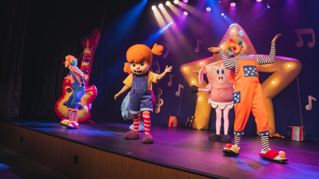 O elenco do espetáculo O Diário de Mika — incluindo a personagem principal, atores caracterizados como palhaços e personagens fantasiadas — apresenta-se no palco iluminado do Teatro Dr. Losso Netto.