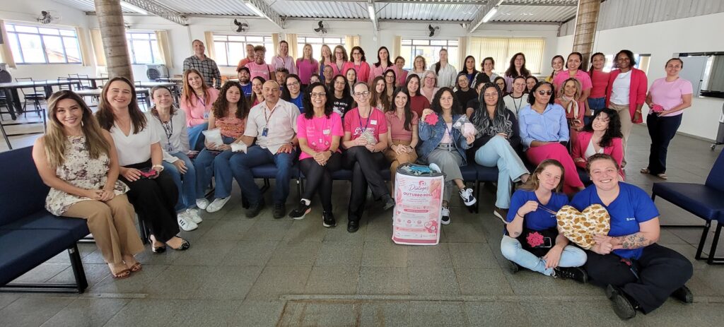 Fotografia coletiva com todos os participantes e organizadores do evento reunidos e a sorrir para a câmara, muitos dos quais vestem peças de roupa cor-de-rosa. Ao centro e em primeiro plano, exibem-se materiais informativos, mimos e almofadas temáticas.