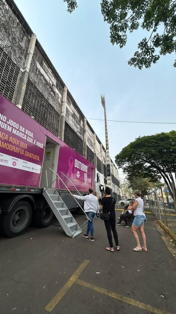 Unidade móvel (camião) da campanha "Outubro Rosa" estacionada junto ao estádio do XV de Piracicaba, com uma escada de acesso e várias mulheres a aguardar atendimento para a realização de mamografias gratuitas.