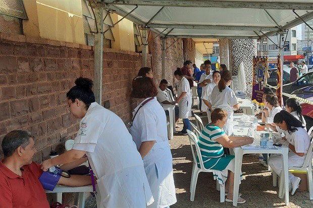 Mostra uma ação comunitária de saúde sob uma tenda ao ar livre ao lado de uma parede de pedra. Profissionais e estudantes de saúde de bata branca realizam exames e atendimentos a várias pessoas sentadas em mesas de plástico, incluindo a afirição de pressão arterial