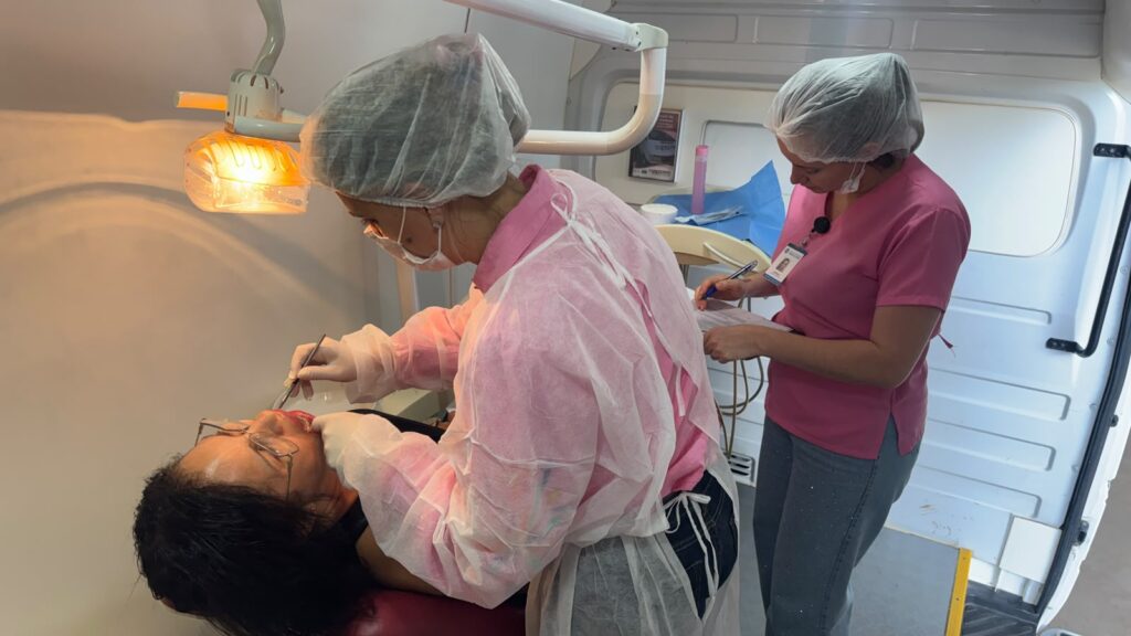 Uma profissional de saúde a realizar um atendimento dentário a uma paciente deitada numa cadeira, enquanto outra auxiliar anota informações clínicas num bloco de notas.