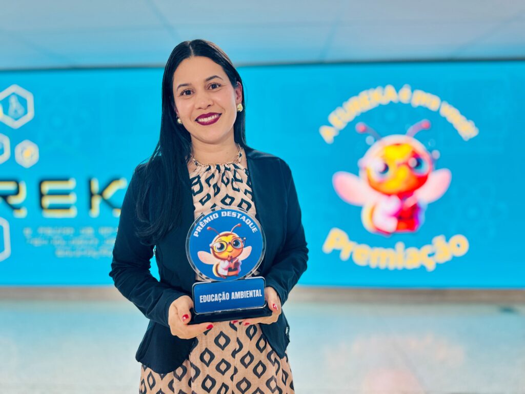 Uma mulher sorri para a câmara enquanto segura um troféu com a inscrição "Educação Ambiental", tendo ao fundo um painel azul com o logótipo da feira Eureka!.