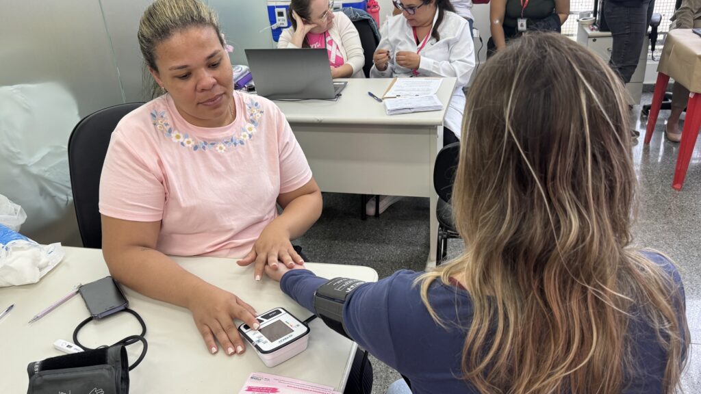 Uma profissional de saúde a realizar o procedimento de aferição de pressão arterial no braço de uma mulher sentada numa mesa.