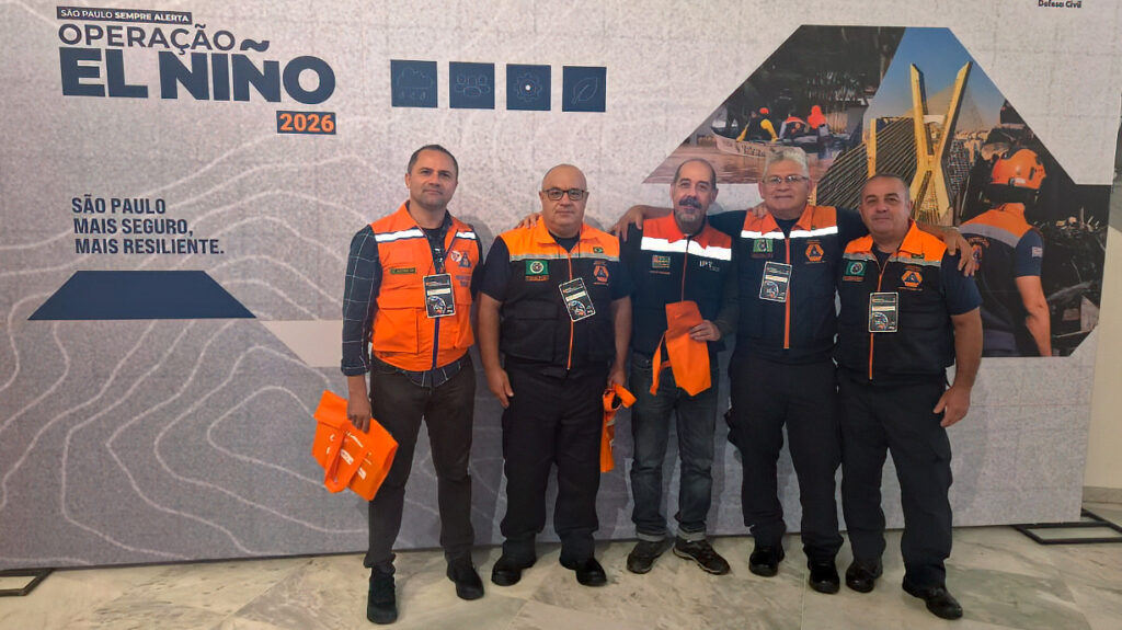 Cinco agentes da Defesa Civil de Piracicaba, devidamente uniformizados, posam para foto em conjunto em frente ao painel oficial do evento "Operação El Niño 2026".