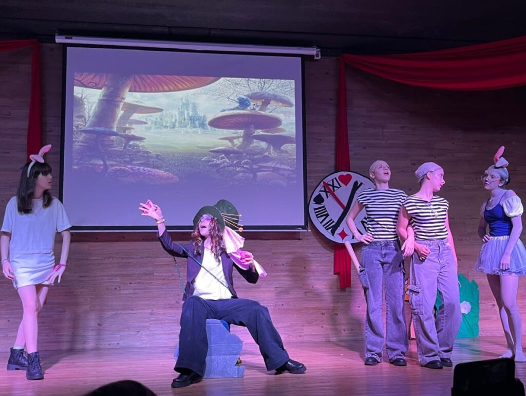 A imagem mostra uma cena de uma peça de teatro escolar inspirada em "Alice no País das Maravilhas", com um grupo de jovens atores em cima de um palco. No centro, um ator encena sentado enquanto gesticula, rodeado por outros atores com figurinos temáticos, tendo ao fundo uma projeção digital e elementos decorativos como tecidos vermelhos e um relógio estilizado.