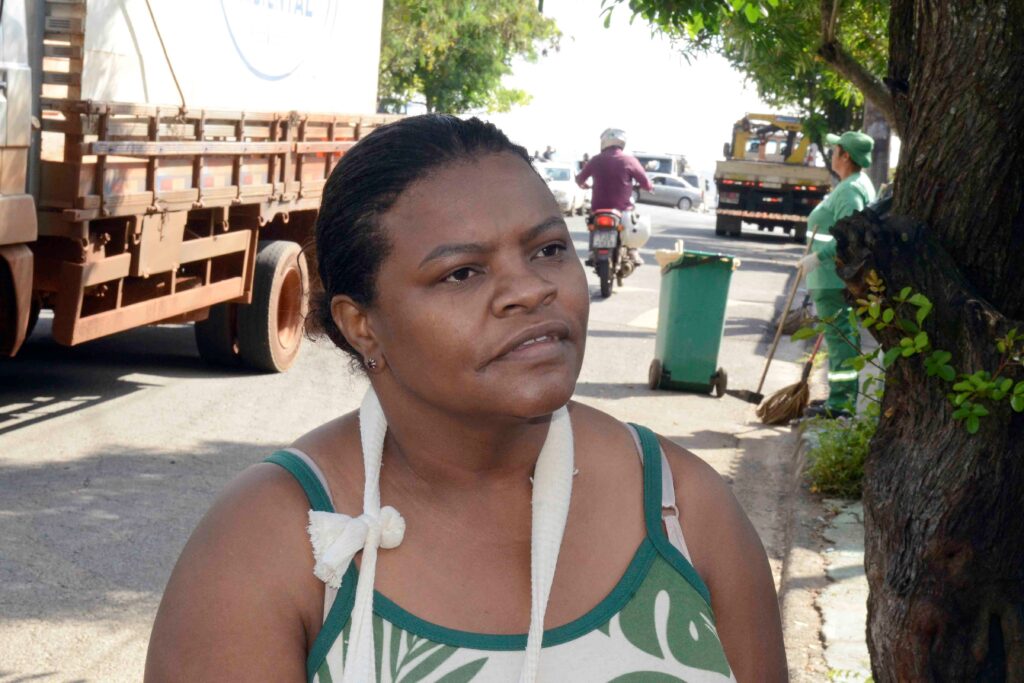 Retrato em plano médio da moradora Aline Aparecida Donato. Ao fundo, vê-se a movimentação da rua, caminhões e uma trabalhadora da limpeza urbanavarrendo a via.