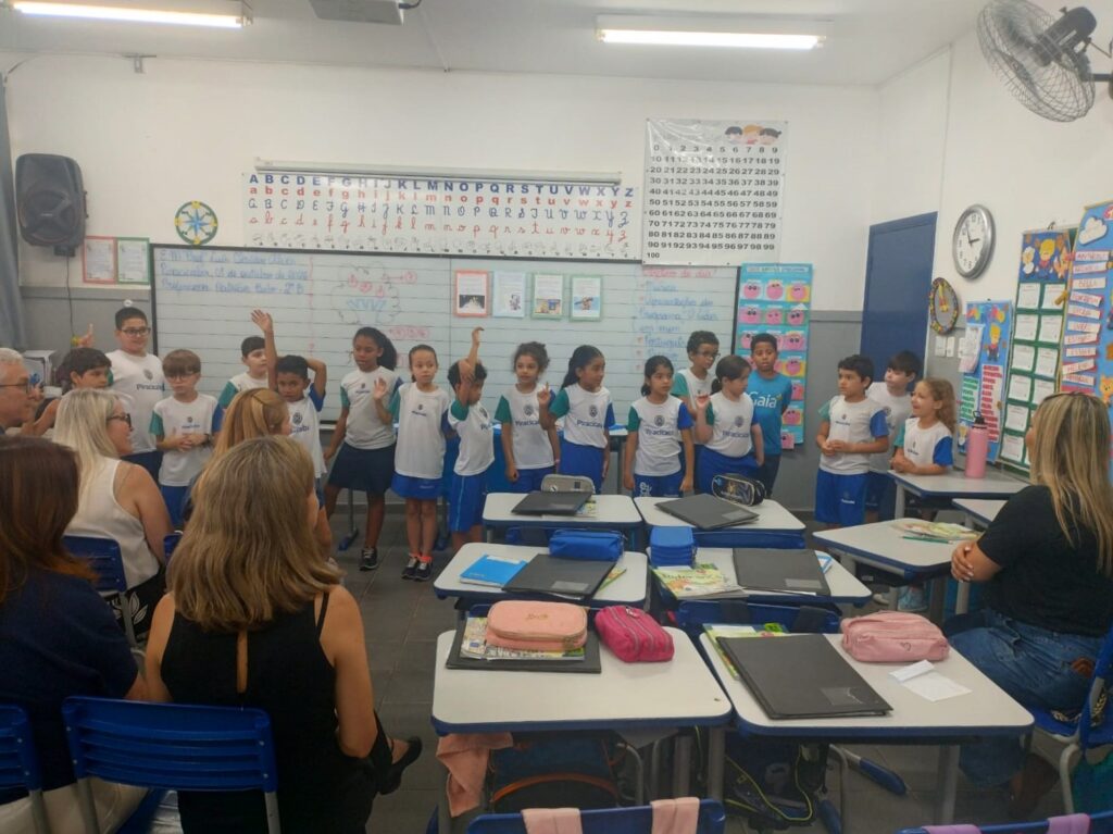 Uma turma de crianças alinhada em frente ao quadro-negro participa numa atividade de sala de aula com os braços levantados, observada por adultos sentados em primeiro plano.