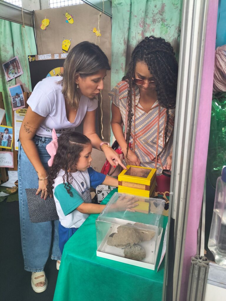 Uma aluna demonstra um experimento a duas adultas que observam atentamente uma caixa amarela e recipientes transparentes sobre a mesa.