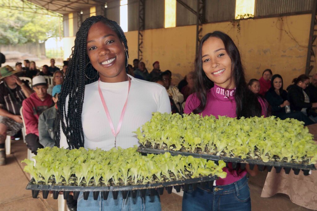 Duas mulheres sorriem para a câmara num espaço coberto enquanto seguram tabuleiros cheios de mudas de alface.