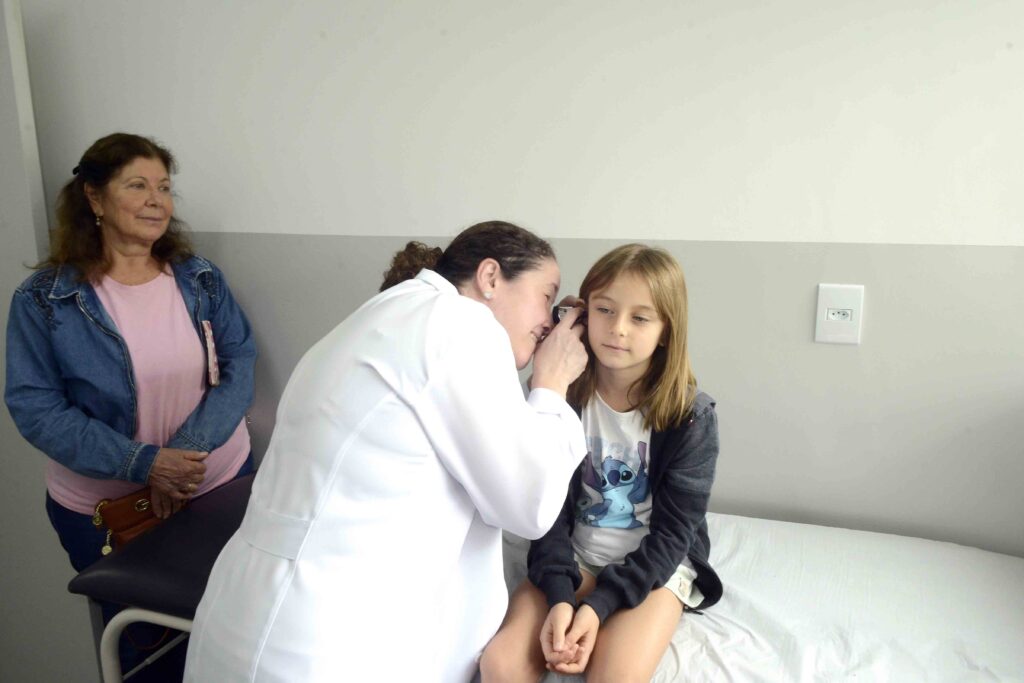 Uma médica de bata branca realiza um exame auditivo com otoscópio a uma criança sentada na marquesa, enquanto uma acompanhante adulta observa ao fundo.