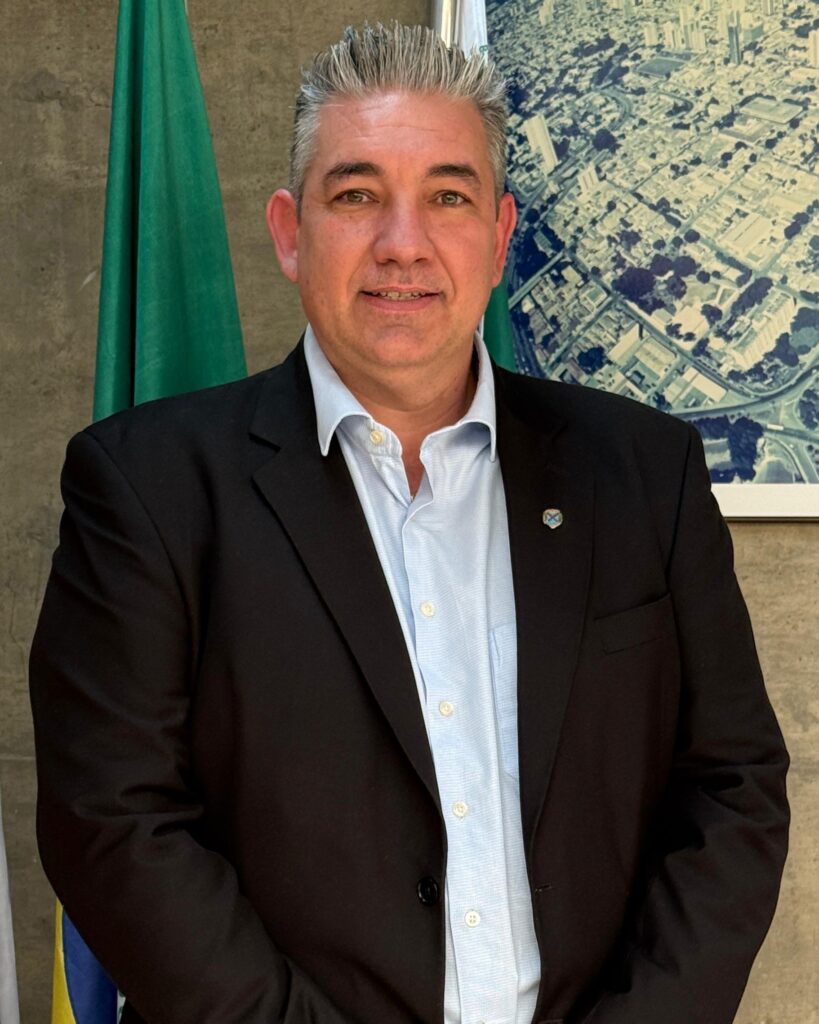 A imagem apresenta um homem de meia-idade vestindo um fato escuro, camisa clara sem gravata e um pequeno crachá/pin na lapela. Encontra-se de pé numa postura formal, posando para uma fotografia oficial, tendo ao fundo bandeiras e um quadro com uma vista aérea urbana na parede.
