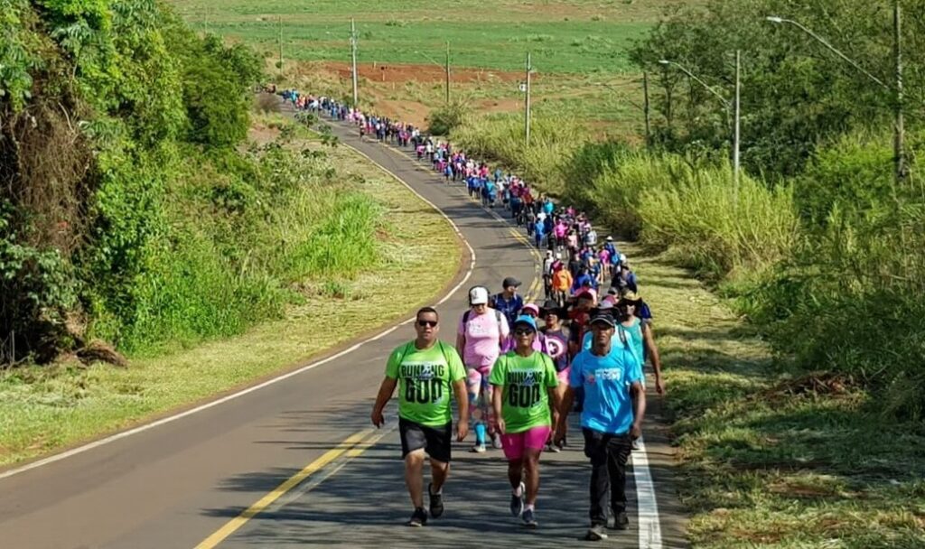 A imagem mostra um grande grupo de pessoas a participar numa caminhada ou corrida ao longo de uma estrada asfaltada que serpenteia por uma zona rural. Os participantes vestem roupas desportivas coloridas e caminham em fila ao longo da via, rodeada por vegetação densa e campos verdes num dia ensolarado.