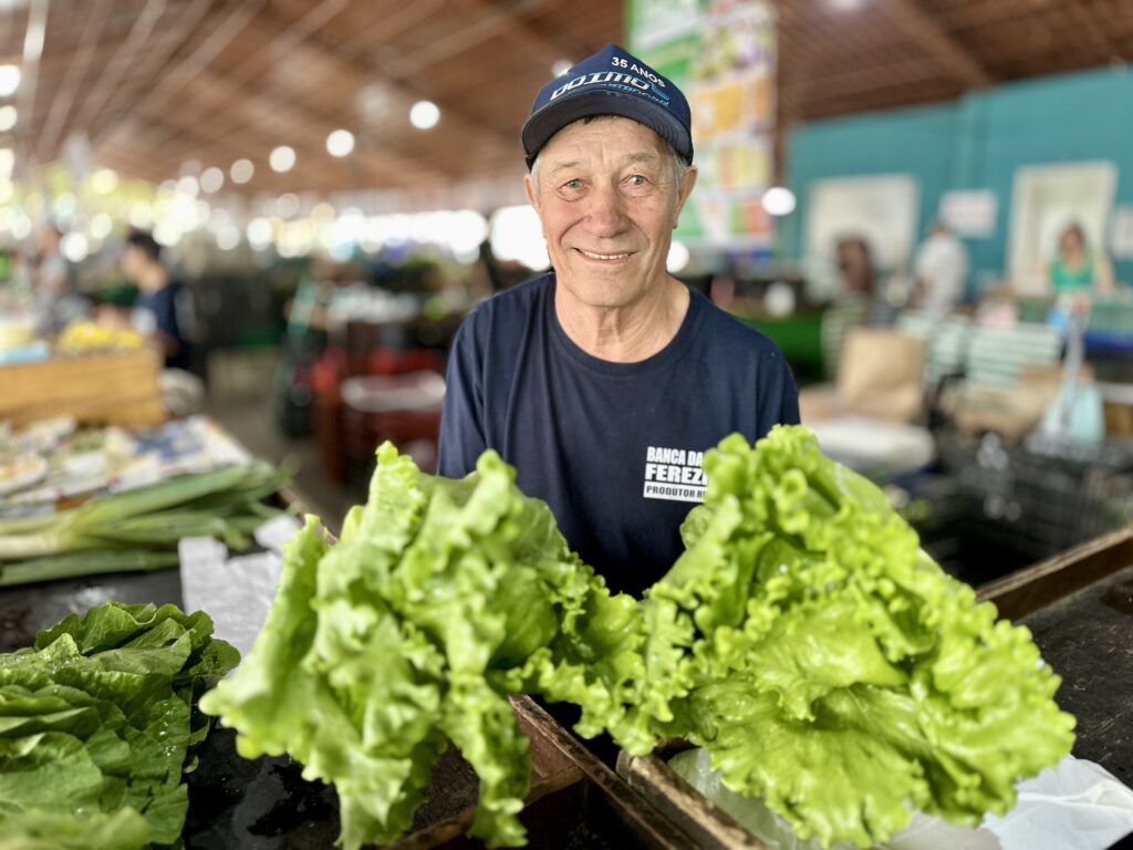 O produtor rural Domingos Ferezin, de 72 anos, sorri atrás da sua bancada de alfaces frescas num varejão municipal.