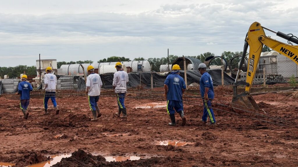 Uma equipa de trabalhadores da construção civil transporta manualmente uma longa armação de ferro sobre um terreno enlameado, ao lado de uma retroescavadora amarela estacionada.