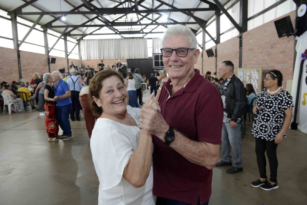 Em primeiro plano, o casal Maria Stella e João Antônio Stella posa sorridente enquanto dança de mãos dadas durante o evento na Estação do Idoso.