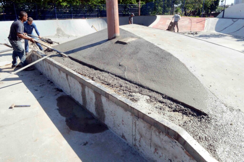 Vista num ângulo mais aberto do mesmo local, mostrando dois trabalhadores em conjunto a alinhar a camada de betão fresco na rampa de transição da pista de skate. O espaço ao redor exibe a estrutura da pista (bowl e caixotes) a ser remodelada.