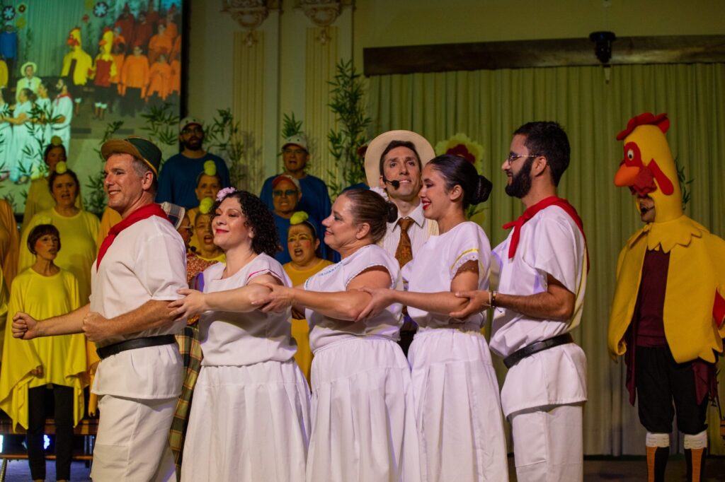 Integrantes do grupo de dança folclórica, vestidos com roupas tradicionais brancas e lenços vermelhos, dançam de mãos dadas à frente do coral e de um figurante caracterizado de galinha no palco.
