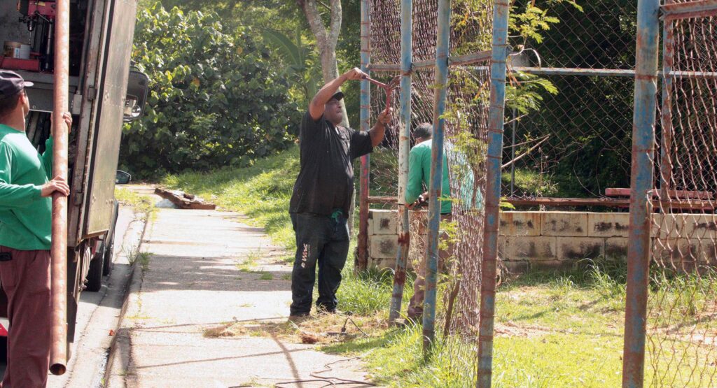 Trabalhadores realizam reformas e ajustes na estrutura metálica da fita/alambrado de uma quadra esportiva comunitária.