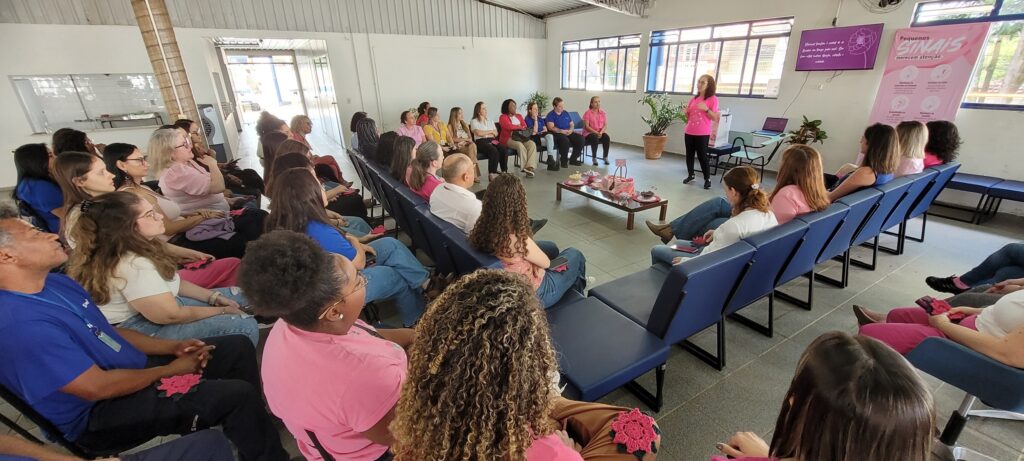 A palestrante, vestindo uma t-shirt rosa e calças escuras, fala de pé no centro da sala a partilhar o seu testemunho perante a plateia compenetrada. Ao fundo, observam-se um ecrã de suporte visual e um painel sobre o Outubro Rosa.