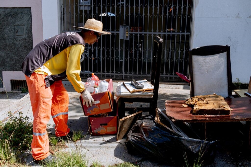 Um agente de limpeza urbana, vestindo chapéu de palha, luvas e fardamento de proteção em tons de amarelo e laranja, recolhe caixas com recipientes e objetos descartados no passeio em frente a uma residência.