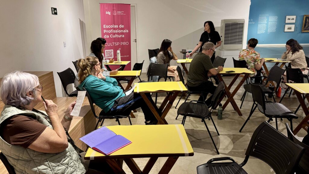 Diversos alunos participam numa oficina de artes plásticas na Pinacoteca (Pina), dispostos em pequenas mesas individuais amarelas enquanto a instrutora orienta a sessão ao fundo.