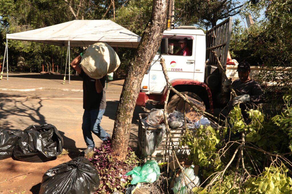 Mostra a equipe de limpeza a recolher grandes sacos de lixo pretos e resíduos acumulados na calçada (como galhos de árvores e plástico) para carregar na caçamba de um caminhão de recolha da EcoTerra. Gostarias de mais informações sobre as zonas afetadas ou sobre as ações da Defesa Civil na região?