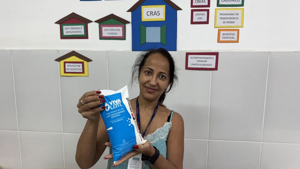 A assistente social Vilmarina Soares segura e exibe um saco de leite pasteurizado do programa Viva Leite em frente a uma parede decorada com quadros informativos dos serviços do CRAS.