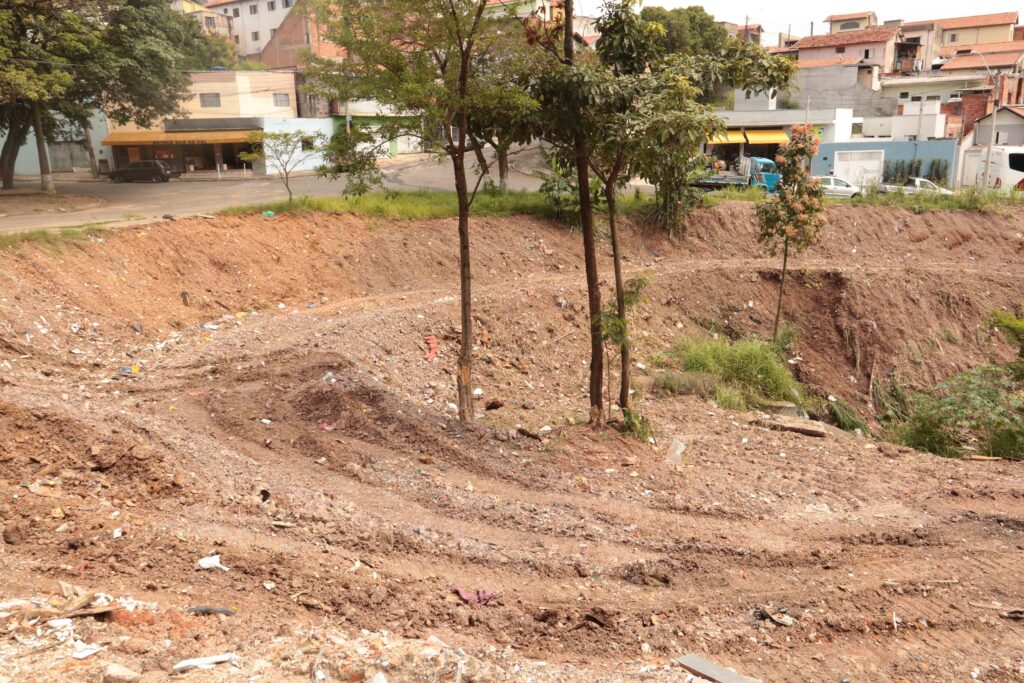 Uma vista ampla de uma encosta de terra já limpa e nivelada por máquinas, mostrando marcas de rastros no solo perto de uma rua urbana.