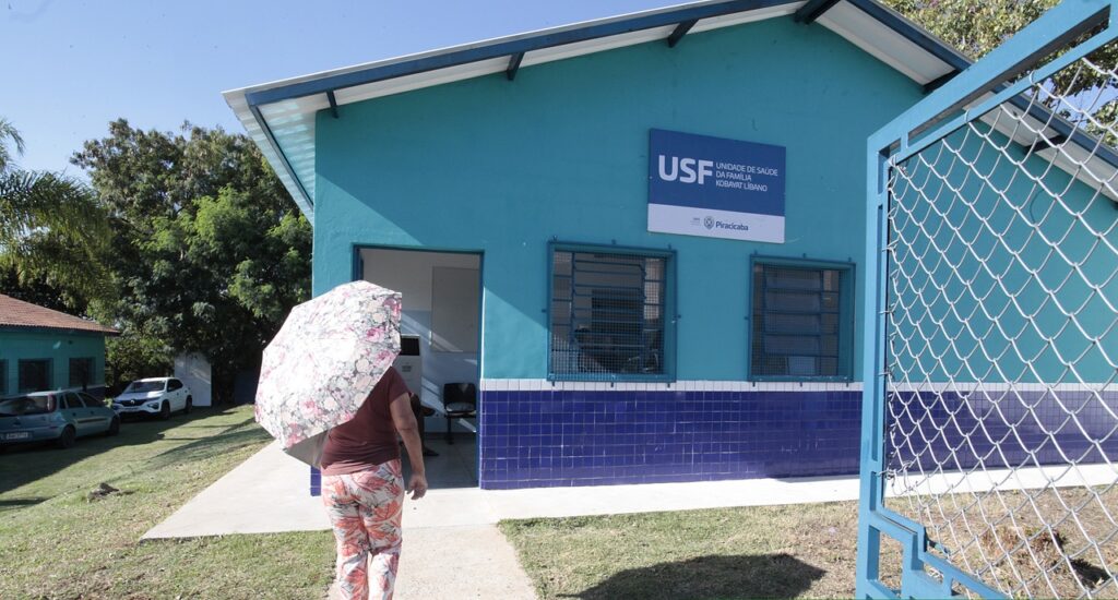 A imagem mostra a entrada de uma Unidade de Saúde da Família (USF), pintada nas cores azul-turquesa e azul-escuro. Há uma pessoa caminhando em direção ao prédio segurando um guarda-chuva florido.