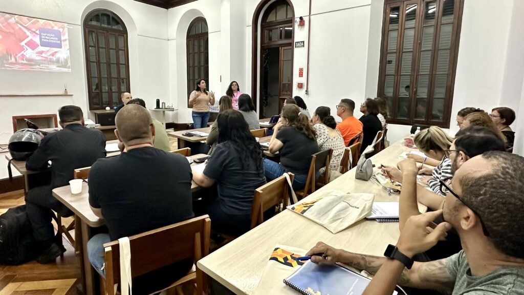 Apresenta uma sala de aula com várias pessoas dispostas em bancadas a prestar atenção a duas instrutoras que falam à frente do espaço. Ao fundo, projeta-se uma apresentação com o título "Captação de Recursos para Eventos".