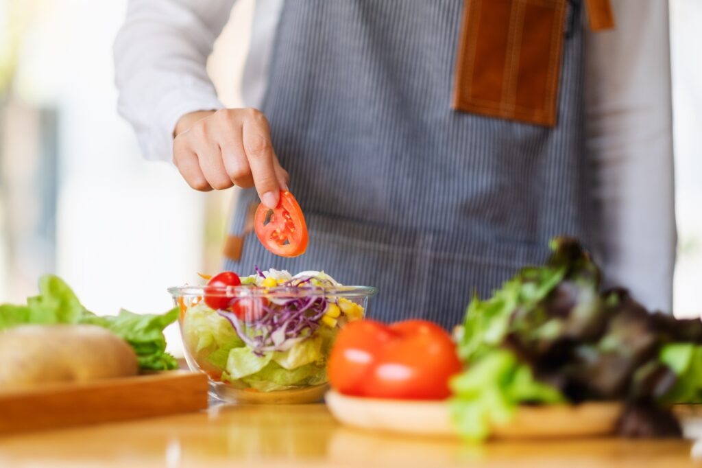 A imagem mostra uma pessoa a preparar uma salada numa bancada de cozinha, vestindo um avental de riscas. Em primeiro plano, a mão segura uma rodela de tomate para colocá-la numa tigela de vidro transparente com alface, repolho roxo e outros ingredientes, rodeada de legumes frescos como tomates inteiros e folhas verdes.