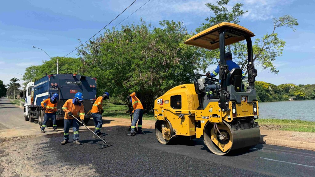 Trabalhadores de uniforme e capacete aplicam e nivelam asfalto fresco numa via urbana perto de um lago, acompanhados por um rolo compressor amarelo da Caterpillar e um camião de suporte ao fundo.