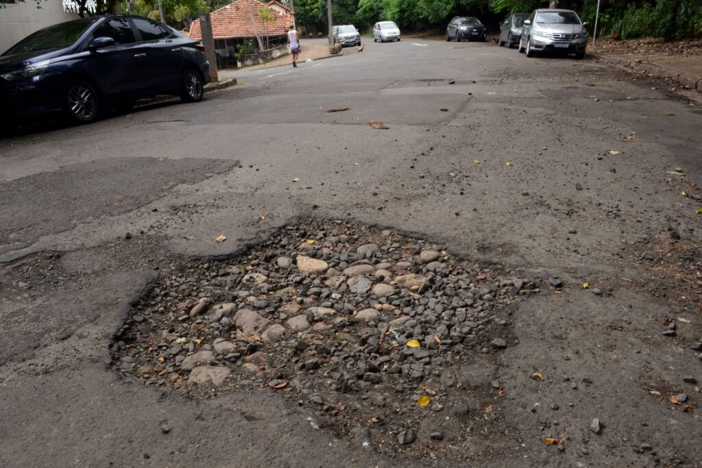 Asfalto deteriorado exibindo uma grande cratera preenchida com pedras e cascalho no meio da via pública.