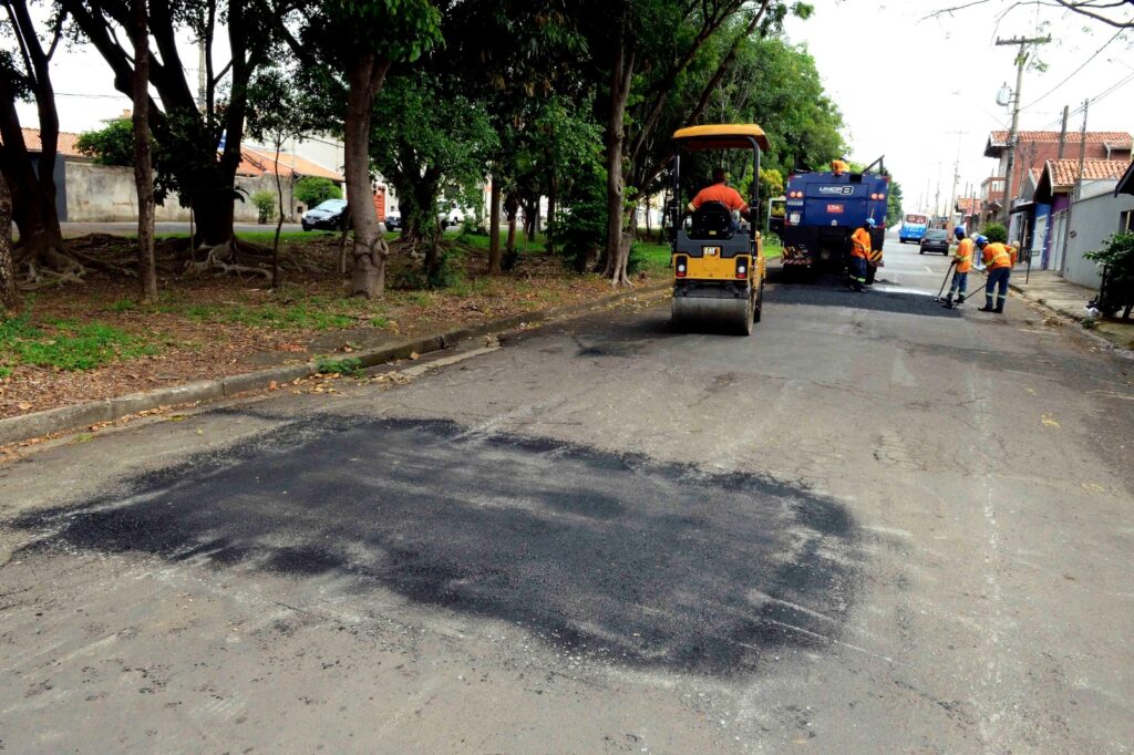 A imagem mostra uma obra de tapa-buracos/recapeamento asfáltico numa rua arborizada. Em primeiro plano, vê-se um trecho de asfalto novo aplicado na via. Mais ao fundo, operadores de uniforme tático/laranja trabalham na pista junto a um camião de pavimentação e a um rolo compactador amarelo.