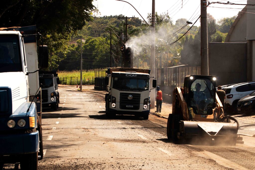 Máquinas pesadas e camiões realizam trabalhos de fresagem e preparação do asfalto numa avenida, levantando poeira e fumaça sob a luz do sol.