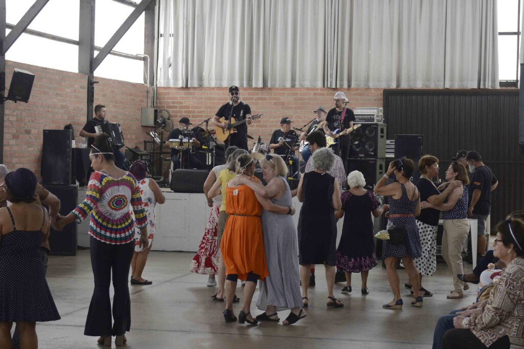 Casais e grupos de idosos dançam no salão ao som de uma banda com sanfona e violão que se apresenta num palco ao fundo.