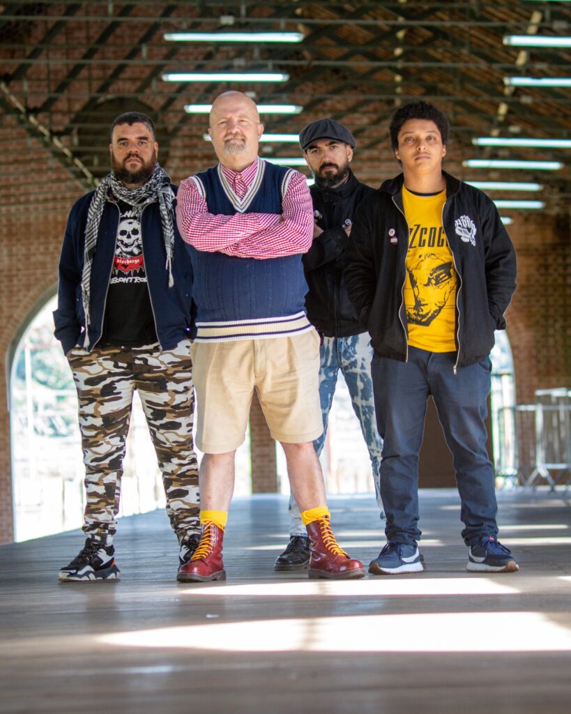 Os quatro integrantes da banda punk Garotos Podres posam alinhados de pé num corredor com estrutura metálica e iluminação no teto.