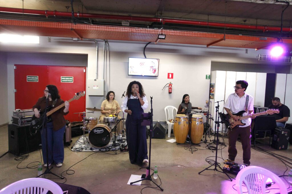 Uma banda musical a atuar ao vivo num espaço interior amplo, com uma vocalista ao centro segurando um microfone e músicos tocando bateria, instrumentos de percussão, baixo e guitarra.