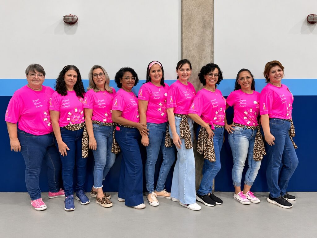 A imagem mostra um grupo de nove mulheres alinhadas lado a lado e a sorrir. Todas vestem camisolas cor-de-rosa associadas à conscientização sobre o cancro da mama (Outubro Rosa) e calças de ganga. Várias delas usam lenços com padrão de leopardo/onça atados à cintura ou como acessório. A fotografia foi tirada num espaço interior com uma parede de fundo branca e azul.