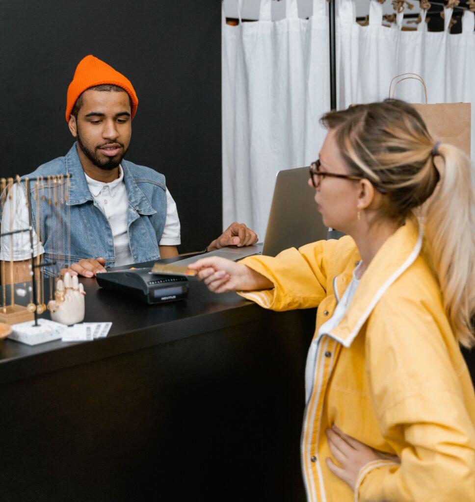 Uma cliente de casaco amarelo e óculos entrega um cartão bancário a um operador de caixa com gorro laranja e colete de ganga para realizar o pagamento na máquina de cartão.