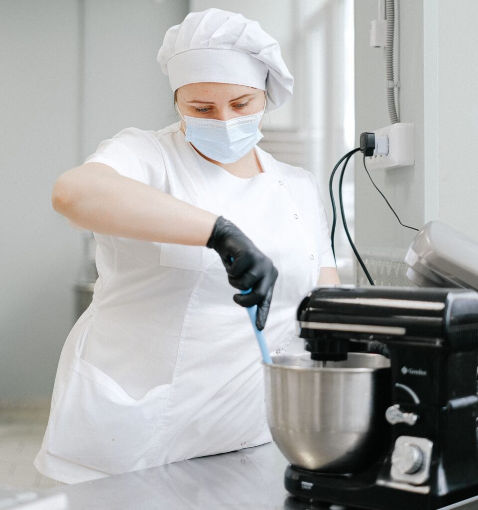 A imagem mostra uma profissional de cozinha vestida com uniforme branco, touca e máscara de proteção, usando luvas pretas enquanto mexe ingredientes numa tigela de batedeira elétrica sobre uma bancada em ambiente de cozinha professional.