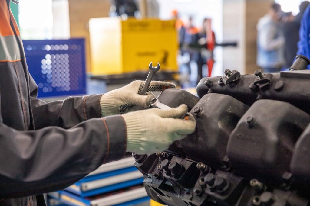 Um mecânico usando luvas de proteção manuseia chaves de fenda e de boca para ajustar peças num motor de grande porte, num ambiente industrial ou oficina.