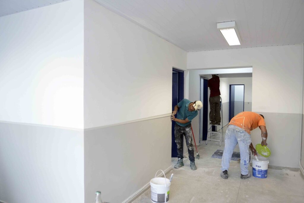 Apresenta três trabalhadores a realizar obras num corredor recém-pintado de branco e cinzento, onde um prepara tinta numa lata, outro ajusta os detalhes do acabamento na parede e um terceiro trabalha sobre um escada ao fundo.